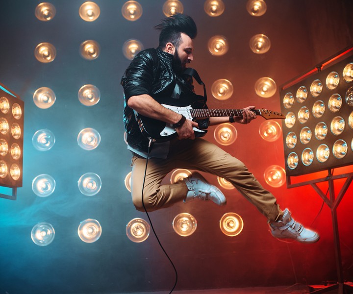 Male performer with electro guitar in a jump on the stage with the decorations of lights. Music entertainment. Bearded musican song performing
